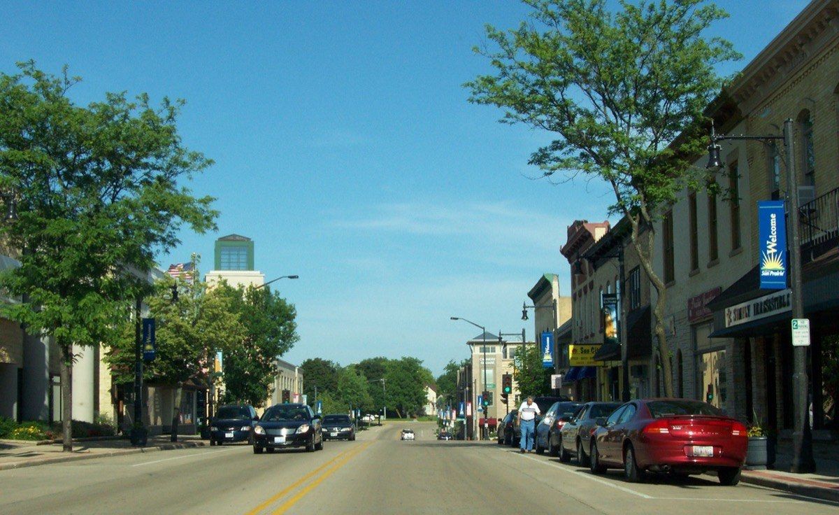 Main Street in Sun Prairie on a clear summer morning, lined with two-story brick storefronts and blue Welcome banners on the lampposts.