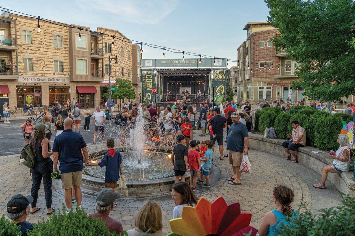 A crowd around the fountain at Cannery Square during the Sweet Corn Festival block party, with a band on stage and Beans 'n Cream Coffeehouse behind.