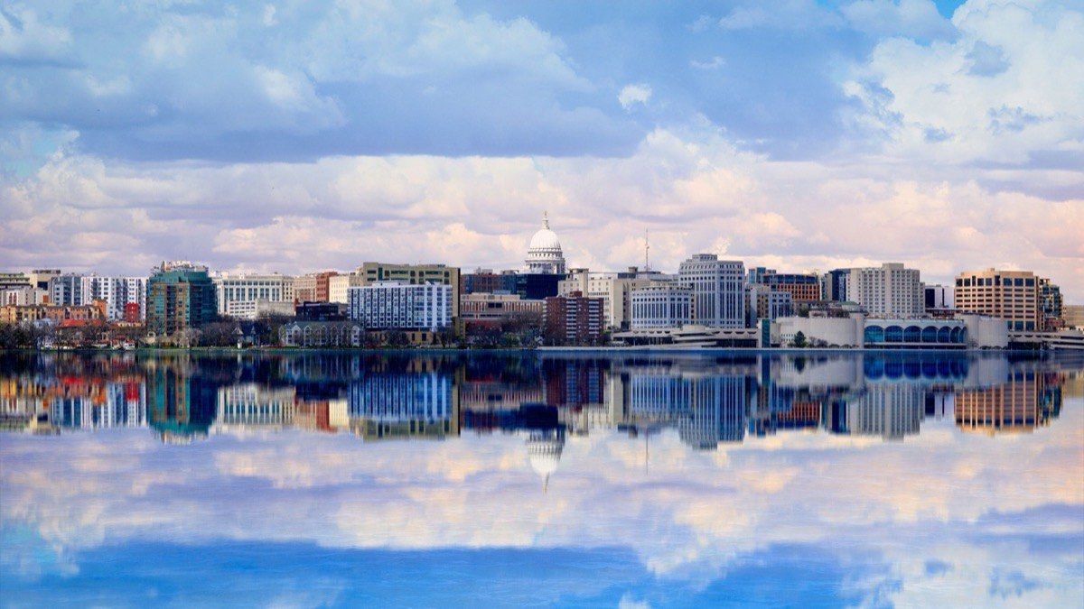 The Madison skyline and State Capitol dome mirrored in the still water of Lake Monona under a cloudy sky.