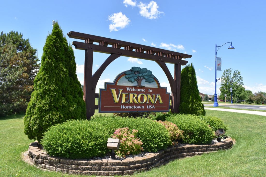 A timber-framed sign reading Welcome To Verona, Hometown USA, surrounded by shrubs and flowers.